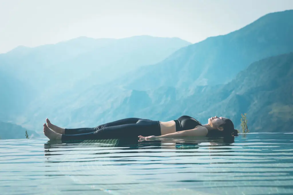 Woman laying in savasana floating on an ocean with mountain backdrop